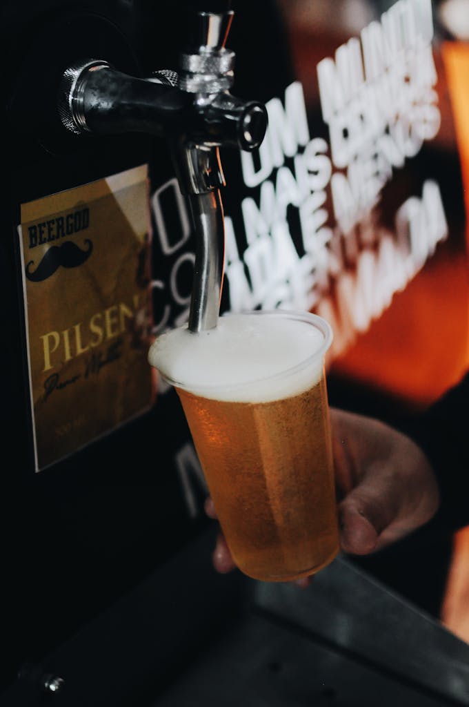 A bartender pours a cold pilsner beer from a tap into a plastic cup, creating a foamy head.