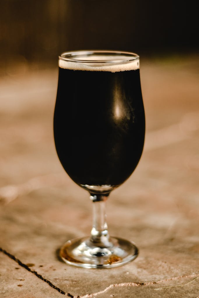 A close-up shot of a dark beer in a tulip glass on a rustic surface.